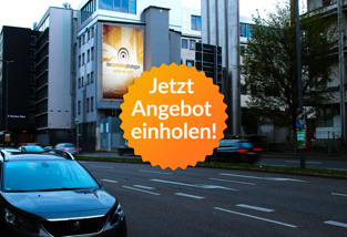 Straßenszene mit mehreren Autos, hohen Gebäuden und einem großen beleuchteten Werbeplakat an der Fassade eines Gebäudes.