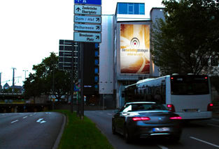Straße mit parkenden Autos, hohen Gebäuden im Hintergrund und einem großen Werbeplakat an der Fassade eines Gebäudes.