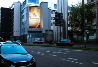 Straße mit Autos und einem Bus, Verkehrsschilder auf der linken Seite, übergroßes beleuchtetes Plakat an einem Gebäude im Hintergrund, Bäume am Straßenrand.